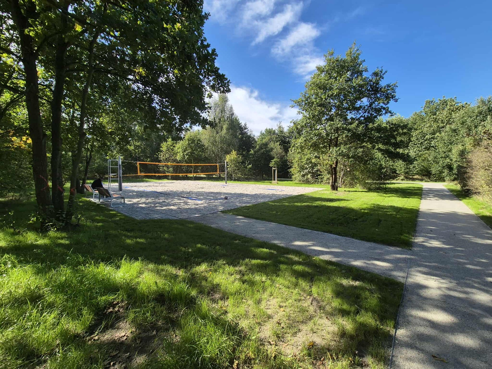 Piaszczyste boisko z siatką i alejka wśród drzew. Boisko do siatkówki plażowej w Parku Maślickim.