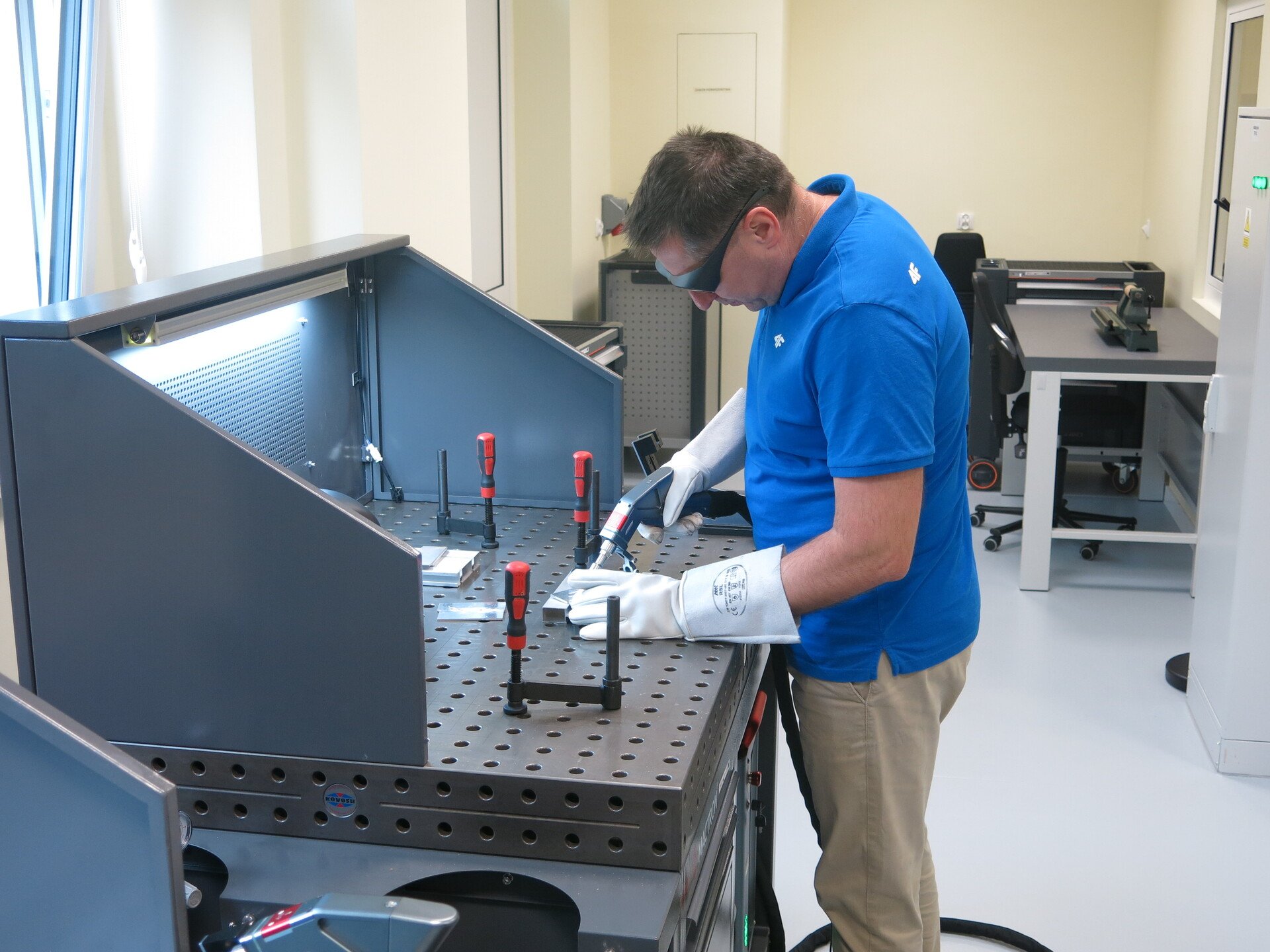 Powiększ obraz: Człowiek w okularach i rękawicach ochronnych  przy stole spawalniczym. Ręczna spawarka laserowa w pracowni BCU przy ul. Strzegomskiej 49a