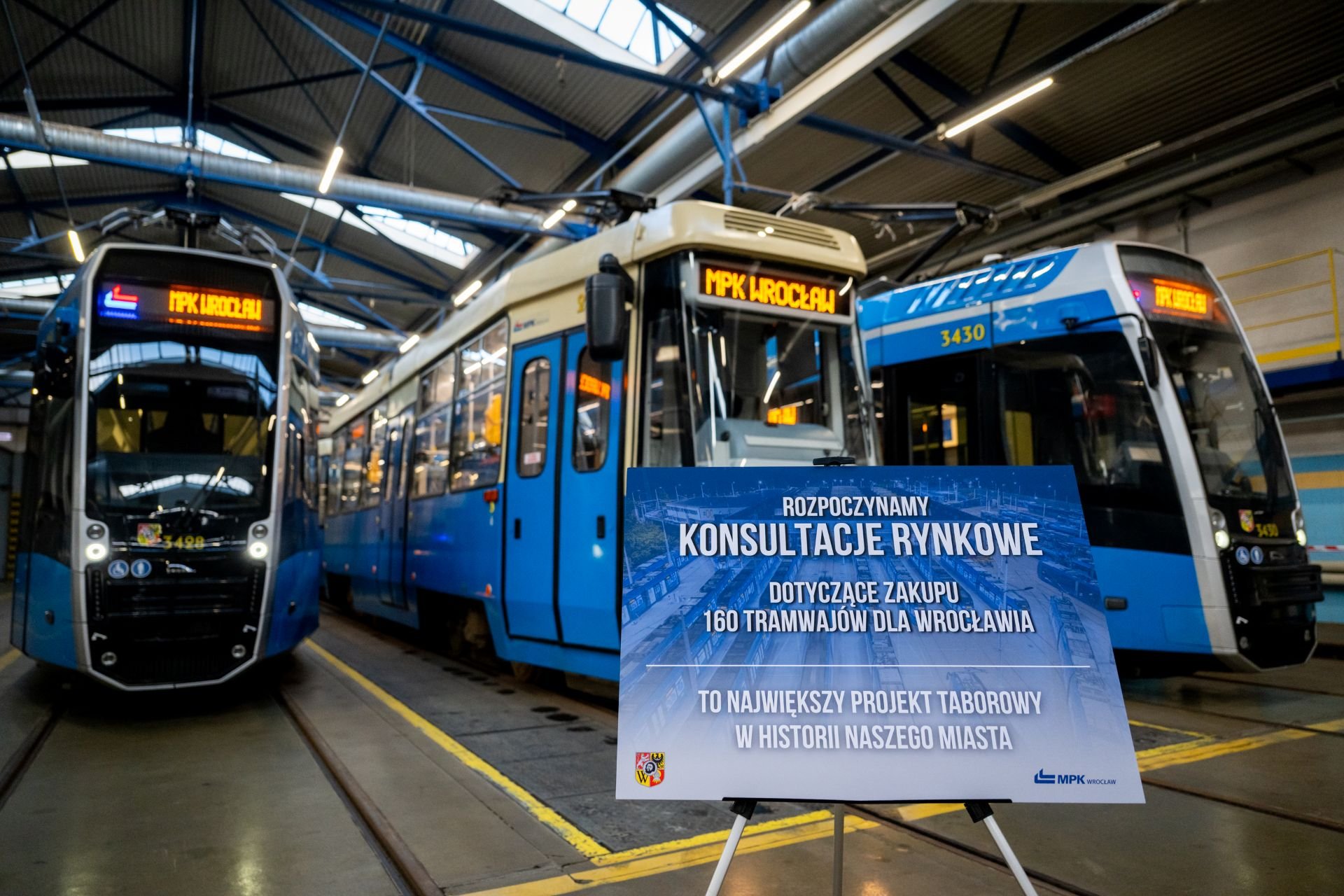 Powiększ obraz: Konferencja MPK Wrocław na zajezdni Borek: tablica z informacją o konsultacjach rynkowych i trzy tramwaje - Konstal 105 i dwie Pesy 146N