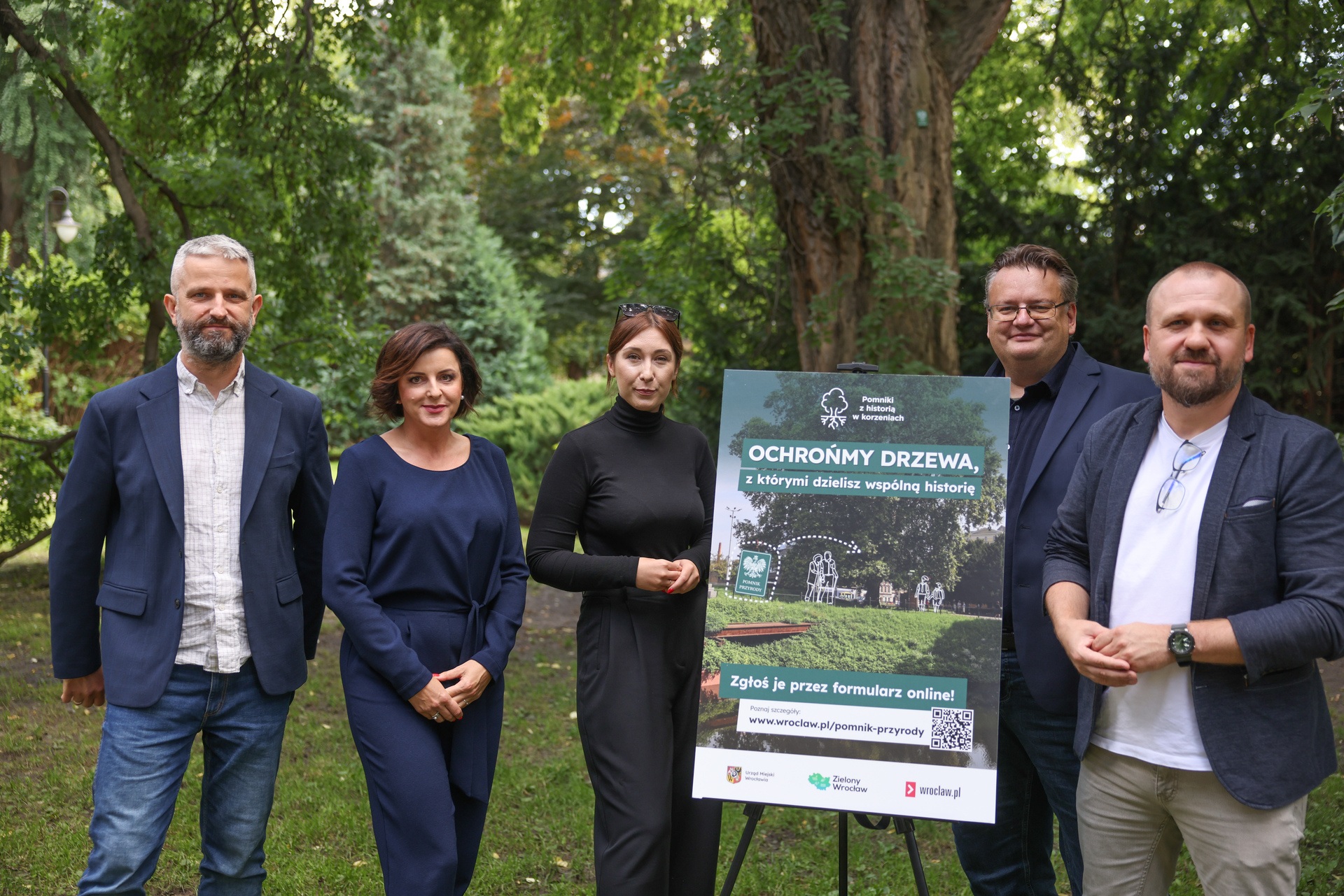 Grupa osób przy tablicy promującej kampanie na tle drzewa w parku. Promocja wrocławskiej akcji „Pomniki z historią w korzeniach”