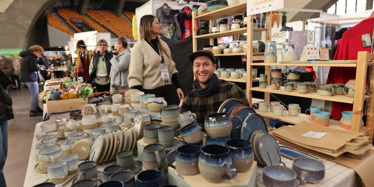 Na zdjęciu, na pierwszym planie, widać ceramikę - kubki i talerze, które są wystawione na sprzedaż na targach w Hali Stulecia. W centralnym miejscu zdjęcia siedzi uśmiechnięty mężczyzna - sprzedawca ceramiki 