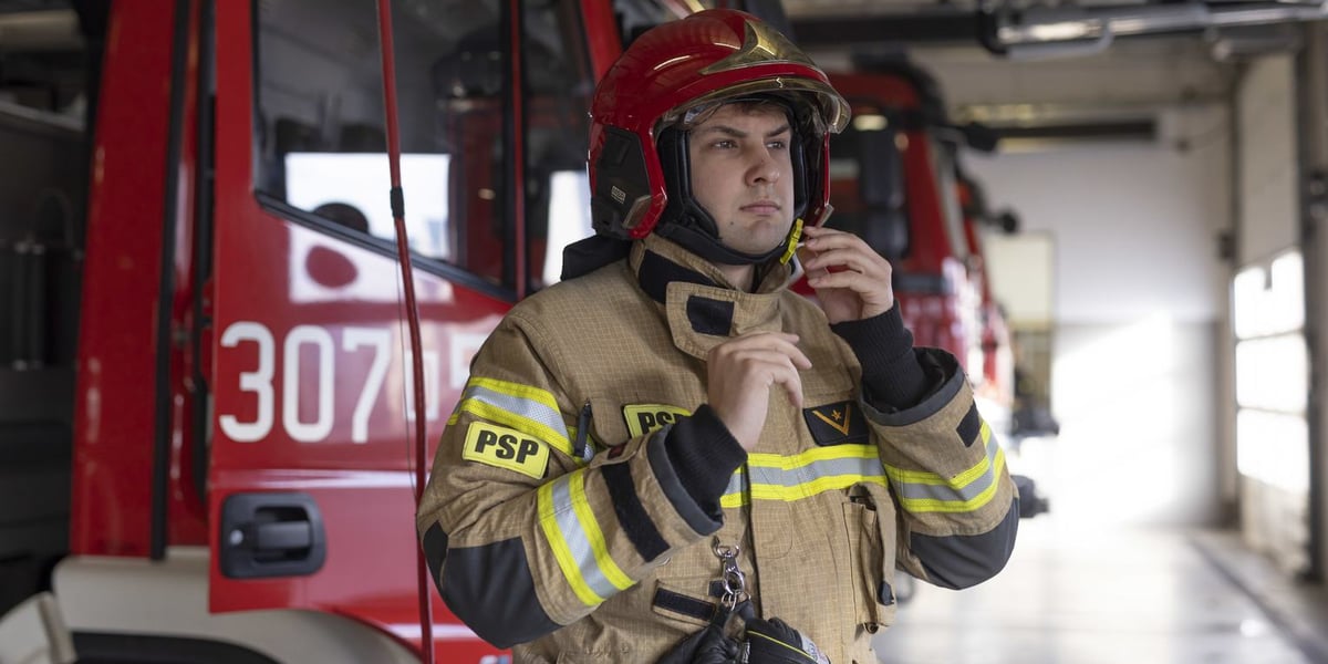 Na zdjęciu st. str. Mateusz Zieliński z Jednostki Ratowniczo-Gaśniczej PSP przy ul. Kosmonautów we Wrocławiu. Mężczyzna jest w strażackim uniformie, w tle widać czerwone wozy strażackie 