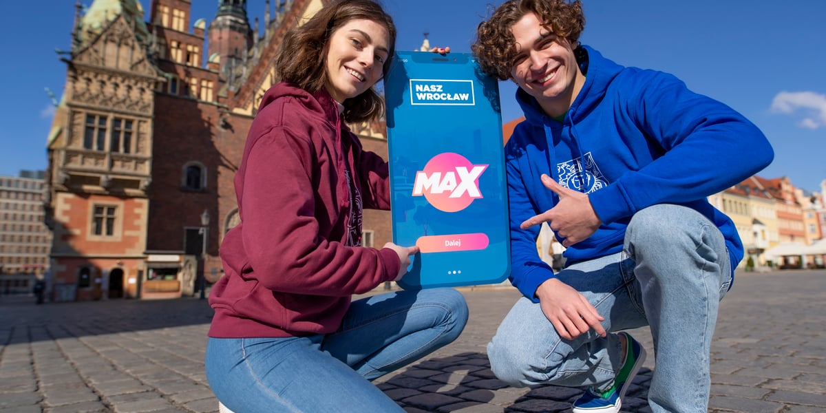 Dwójka młodych ludzi na tle wrocławskiego Ratusza, pomiędzy nimi plakat imitujący smartfona, promujący program Nasz Wrocław Max