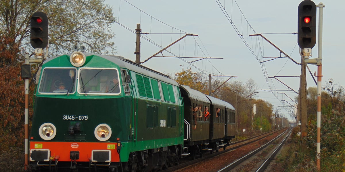 Historyczny pociąg z wagonami jedzie na torach, z okien wychylają się pasażerowie
