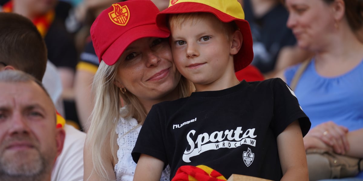 Na trybunach stadionu matka w czerwonej czapce z daszkiem i siedzącym na jej kolanach synkiem w koszulce Betardu i żółto-czerwonym kapelusiku 