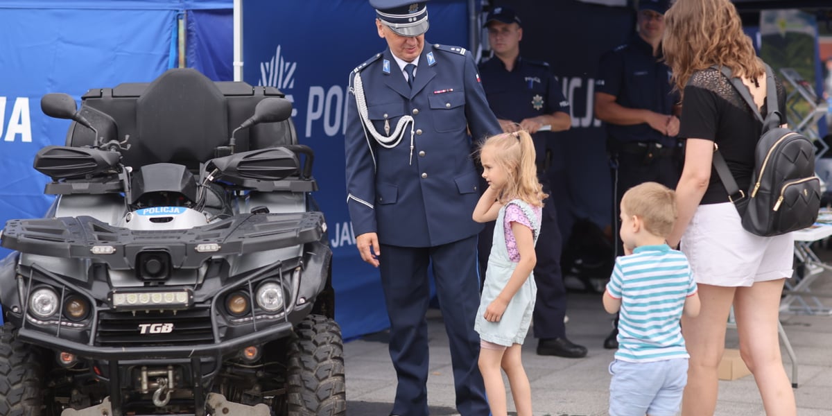 Po lewej pojazd policyjny, obok stoi umundurowany policjant, który patrzy na stojącą obok dziewczynkę. Po prawej tyłem stoi kobieta i chłopiec