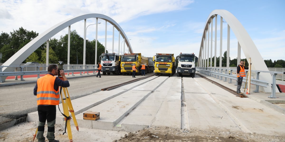 Technik w pomarańczowej kamizelce patrzy przez przyrząd pomiarowy na ciężarówki stojące na moście Chrobrego