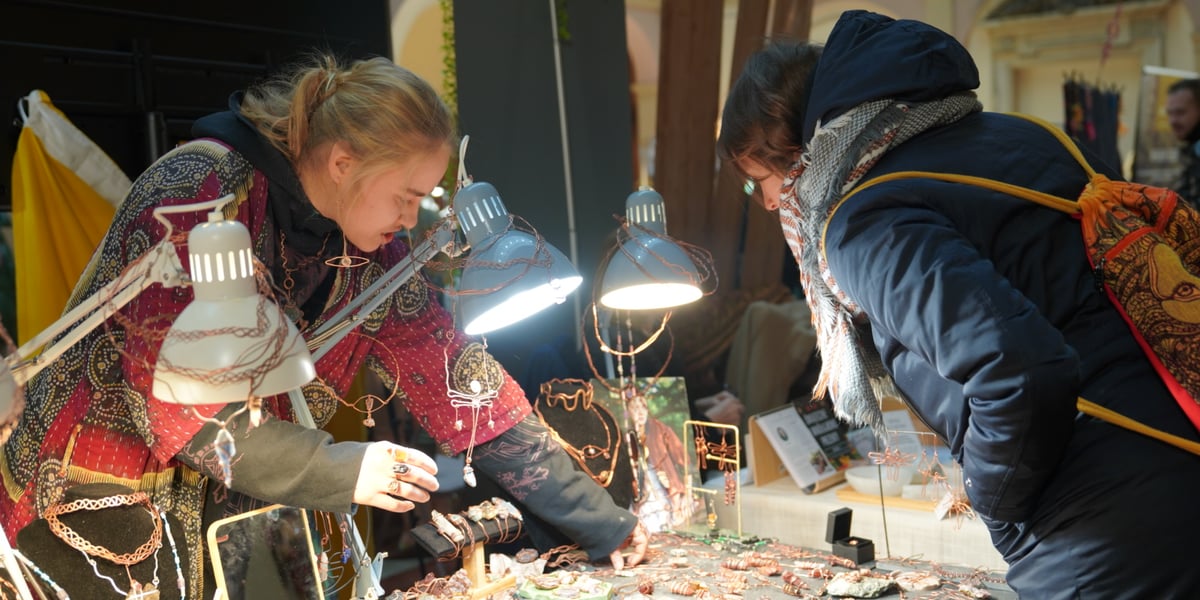 Stoisko z biżuterią artystyczną. Sprzedawczyni w czerwonej bluzce pokazuje towar klientce ubranej w kurkę; Targi ezoteryczne 2 grudnia 2023 