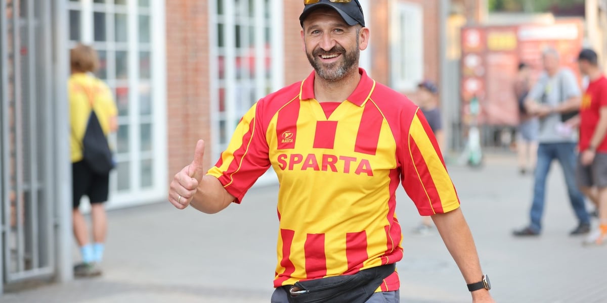 Uśmiechnięty mężczyzna, unoszący kciuk, w żółto-czerwonej koszulce Sparty przed stadionem 