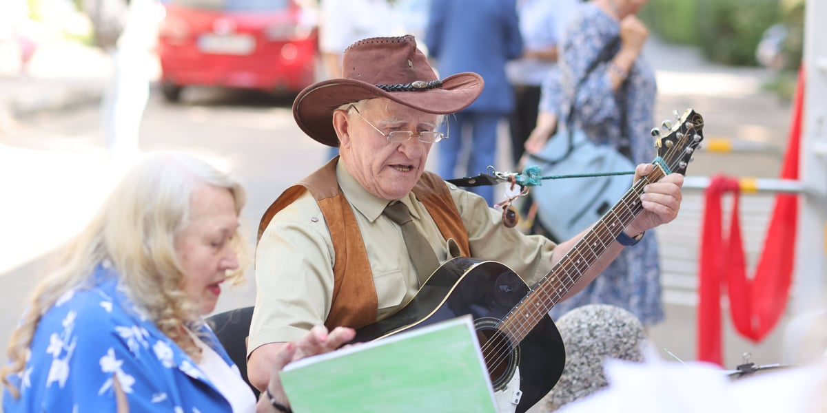 Mężczyzna w kapeluszu z szerokim rondem gra na gitarze, obok śpiewa kobieta w niebieskiej sukni. Otwarcie Centrum Aktywności Lokalnej na Hubach (31.07.2024)