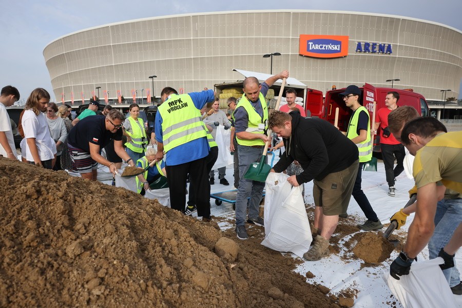 Grupa osób wokół hałdy piachu, sypią go do worków, za nimi widać stadion