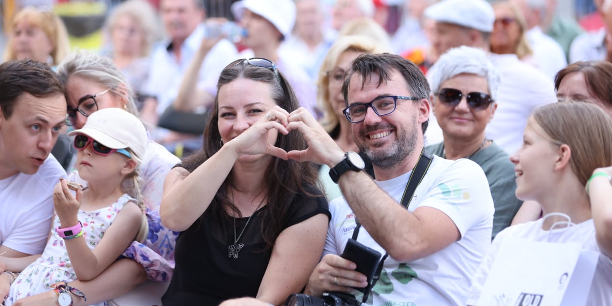 Publiczność siedząca na krzesełkach w Rynku na Old Jazz Days. Na pierwszym planie kobieta i mężczyzna w okularach, uśmiechają się i układają dłonie w kształt serca