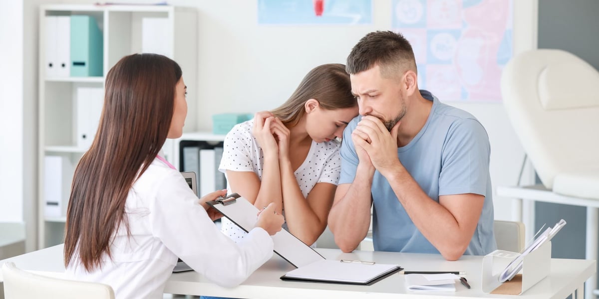 Zdjęcie ilustracyjne - zasmucona młoda para siedzi w gabinecie u lekarki pokazującej wyniki badań.