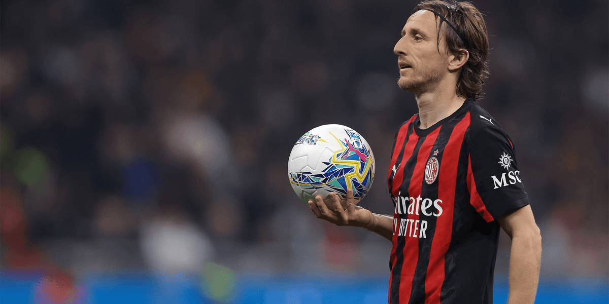 Luka Modrić w czarno-czerwonym stroju Milanu, trzymający piłkę na dłoni
