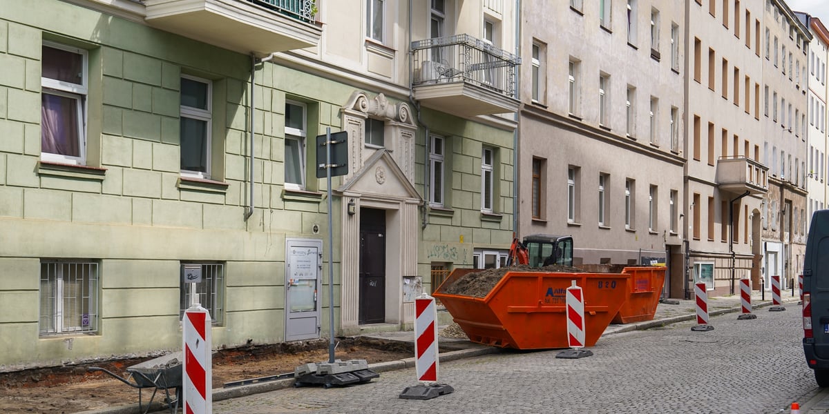 Słupki drogowe i kontenery na gruz stojące na brukowanej jezdni Pl. Konstytucji 3 Maja przy krawędzi chodnika, który jest remontowany.