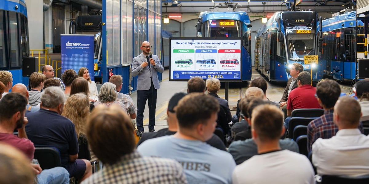 Prezes MPK Wrocław Witold Woźny na spotkaniu z mieszkańcami. W tle widoczne są różne modele tramwajów, koło prezesa stoi tablica z tytułem "Ile kosztuje dziś nowoczesny tramwaj?"