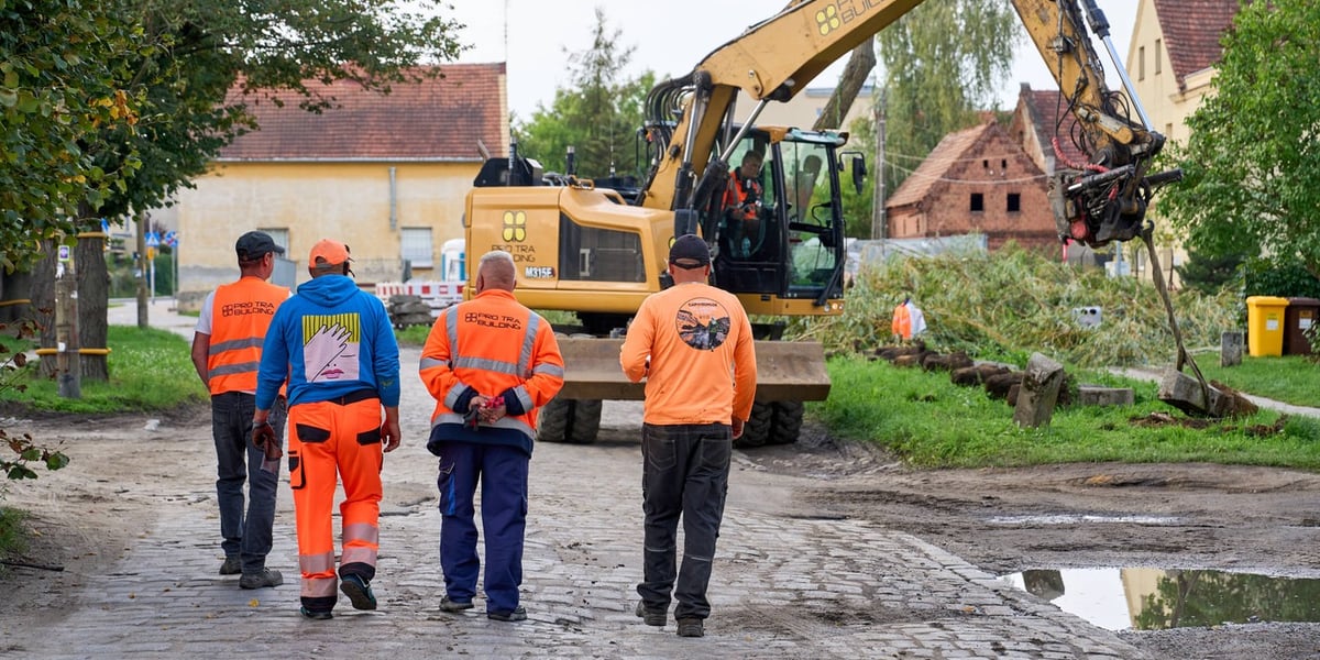 Czterej robotnicy w jaskrawych, pomarańczowych i niebieskich ubraniach idą brukowaną drogą w stronę dużej, żółtej koparki. W tle widać domy i drzewa. Ulica Pawia od soboty jest zamknięta.