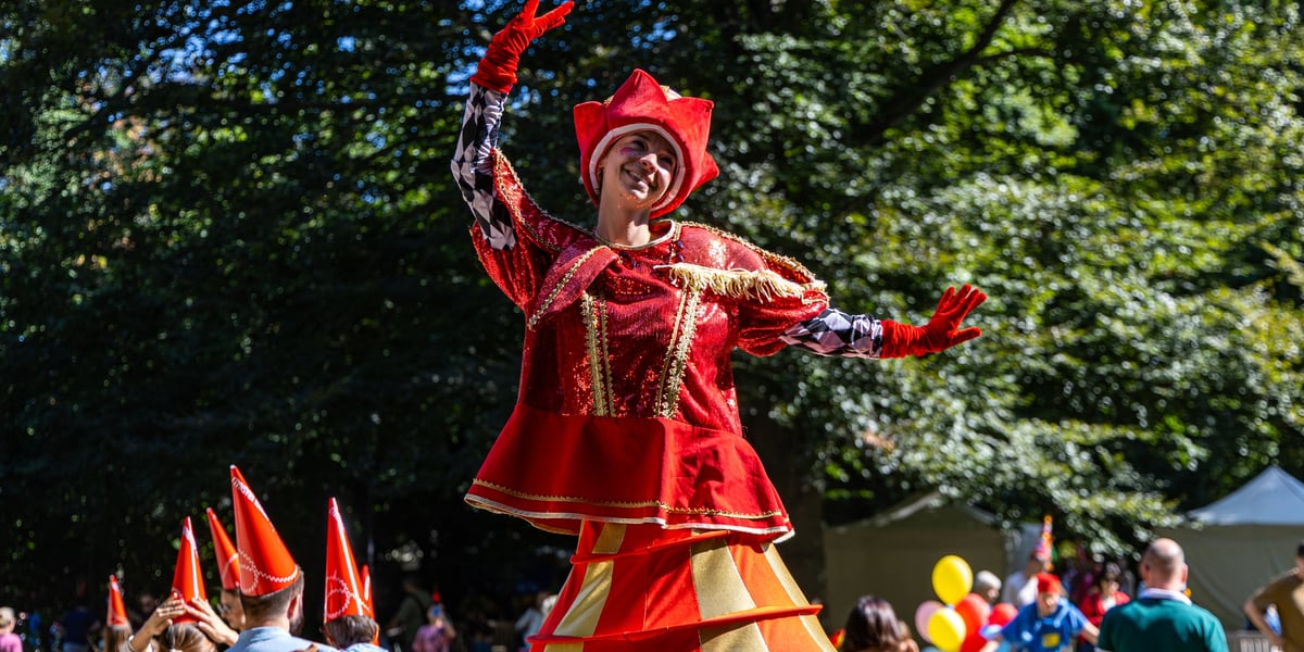 Uśmiechnięta animatorka na szczudłach, ubrana w czerwono-złoty kostium, pozuje z uniesionymi rękami ponad tłumem uczestników Festiwalu Krasnoludków w parku. Zakończył się 17. Wrocławski Festiwal Krasnoludków, 12.09.2026