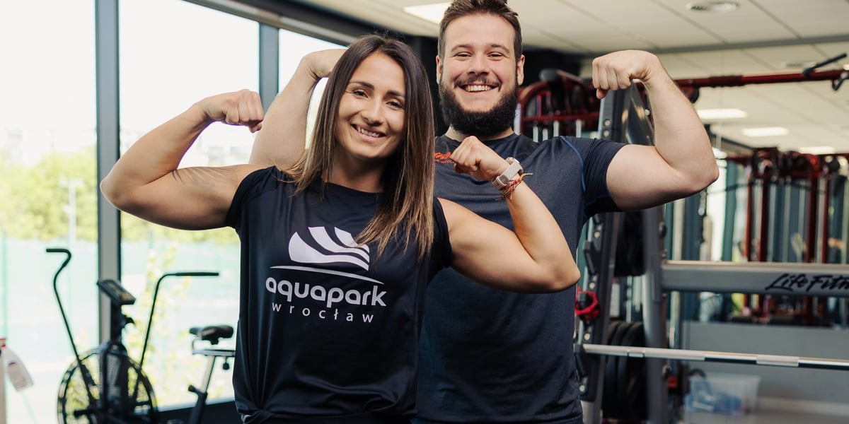 Uśmiechnięta kobieta w koszulce z logo Aquaparku Wrocław i mężczyzna z brodą prężą bicepsy na siłowni. Darmowy e-book Aquaparku Wrocław: trening, dieta i regeneracja