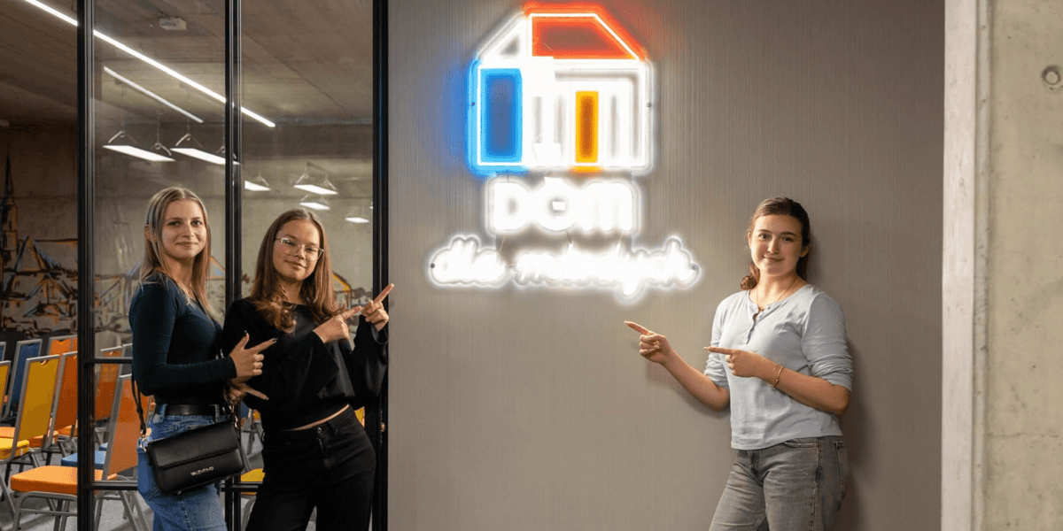 trzy młode dziewczyny wskazują na podświetlany logotyp z napisem "Dom dla Młodych"