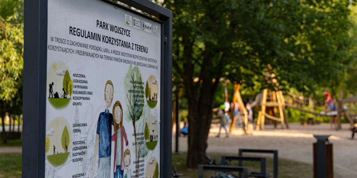 Park Wojszycki. Na pierwszym planie tablica z regulaminem, w tle plac zabaw.