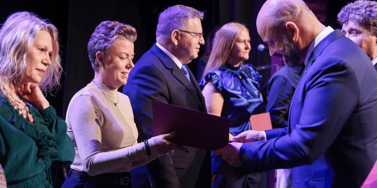 Kobieta w okularach i ciemnej sukience ściska dłoń mężczyzny w granatowym garniturze, który wręcza jej bordową teczkę podczas uroczystości na scenie, w tle stoją inne osoby. Wrocław nagradza nauczycieli i dyrektorów. 224 osoby z Nagrodą Prezydenta Miasta w 2026 roku. 