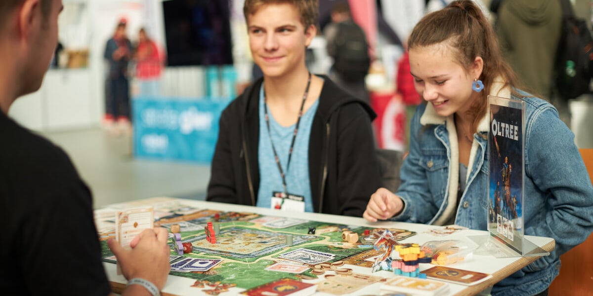 Wrocław Games Week. Wiele z gier planszowych pokazywanych w Hali Stulecia, można wypróbować na miejscu. Na zdjęciu widać trójkę graczy siedzących przy stoliku z grą planszową.