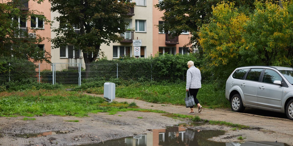 Na zdjęciu starsza pani idąca ścieżką w stronę bloku, na pierwszym planie olbrzymia kałuża. Podwórko w kwartale ulic: Pereca, Szczęśliwej, Jemiołowej i Lwowskiej.