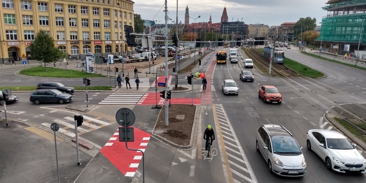 Na zdjęciu dwie trasy dla rowerów, od skrzyżowania ulic - Legnickiej z Rybacką, do przejścia podziemnego pod pl. Jana Pawła II. Po lewej nowa droga rowerowa, po prawej wydzielony pas dla jednośladów na jezdni, po którym jedzie dwóch rowerzystów.