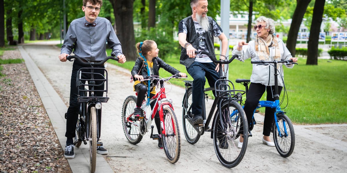 Na zdjęciu grupa rowerzystów w parku, dwóch mężczyzn, dziecko i starsza kobieta 