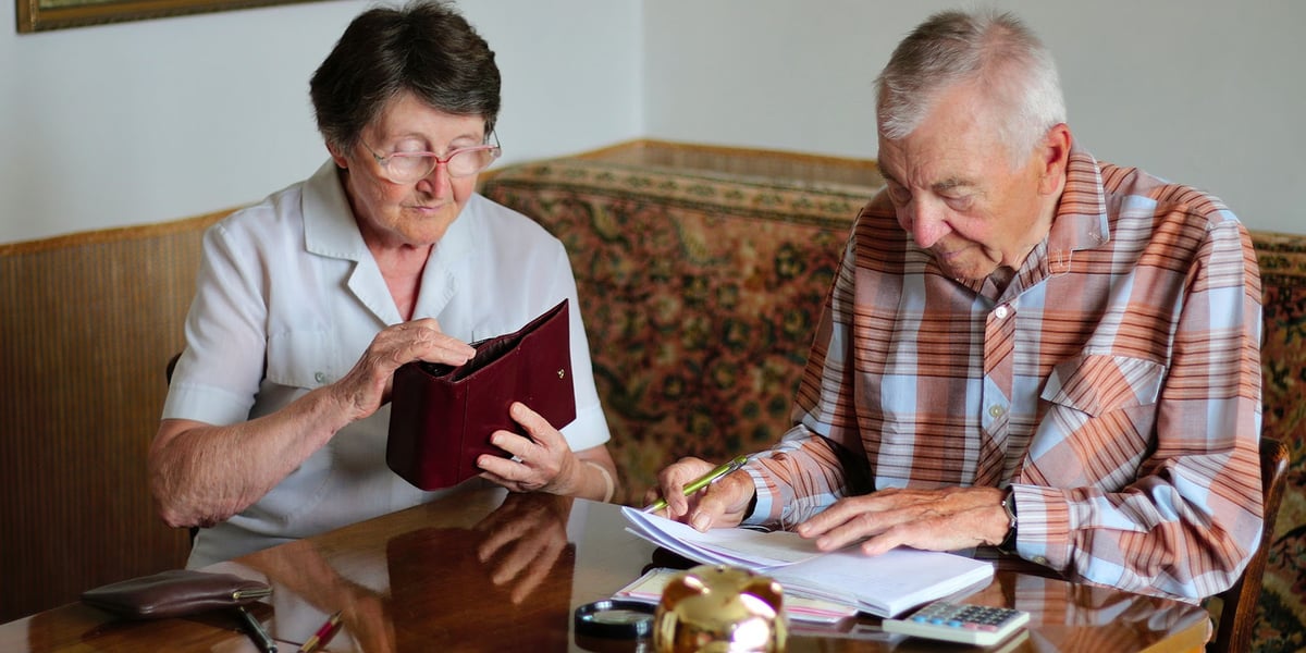Na zdjęciu widać seniorów: kobietę i mężczyznę siedzących przy stole, na którym leżą pieniądze i dokumenty 
