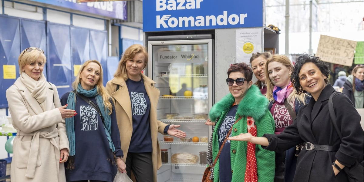 Otwarcie jadłodzielni na Bazarze Komandor. Na zdjęciu widać siedem uśmiechniętych kobiet, stojących po bokach nowej lodówki do dzielenia się produktami spożywczymi.