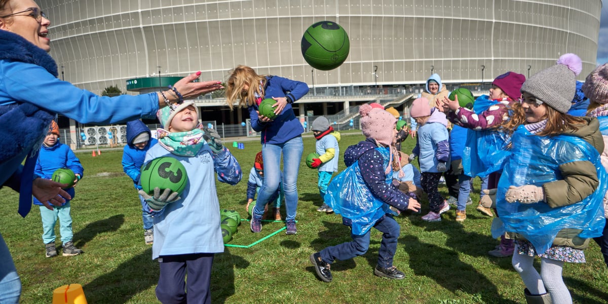 Dzieci i opiekunki bawią się piłką i skaczą, w tle stadion
