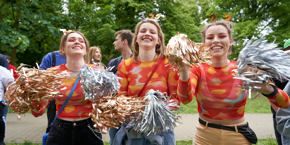 Trzy uśmiechnięte cheerleaderki ubrane w pomarańczowe bluzki, w dłoniach trzymają złote i srebrne pomponami z folii, a na głowie mają opaski z literami T 22