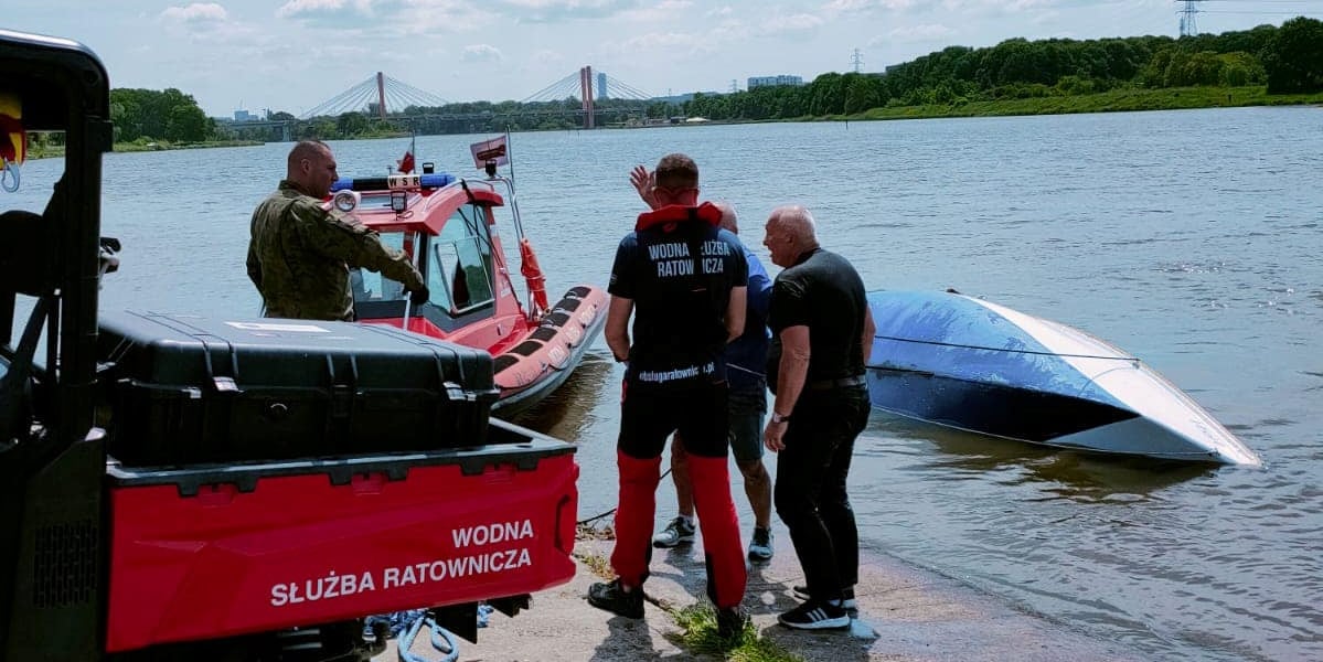 Ratownicy Wodnej Służby Ratowniczej na brzegu, na wodzie widać przewróconą do góry dnem łódź