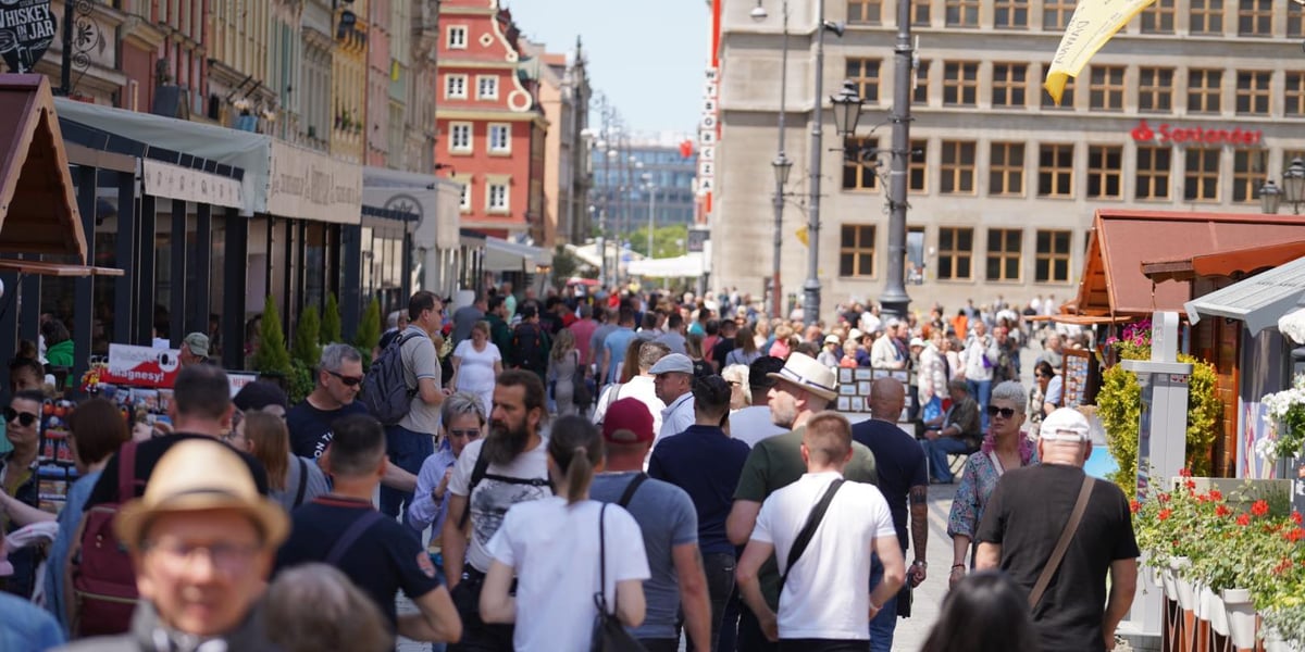 Tłum ludzi chodzących między drewnianymi straganami na Jarmarku Świętojańskim