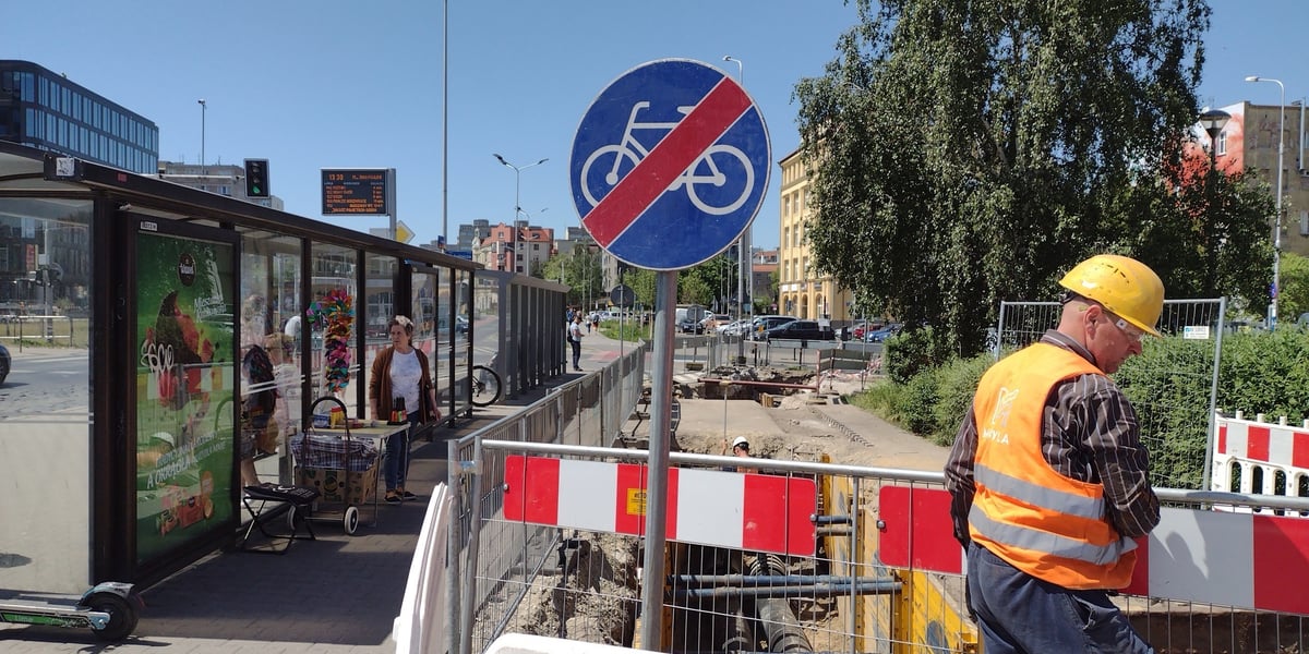 Rów w drodze rowerowej, przekreślony znak z symbolem roweru, mężczyzna w kamizelce i kasku, a także tylna strona wiaty przystankowej