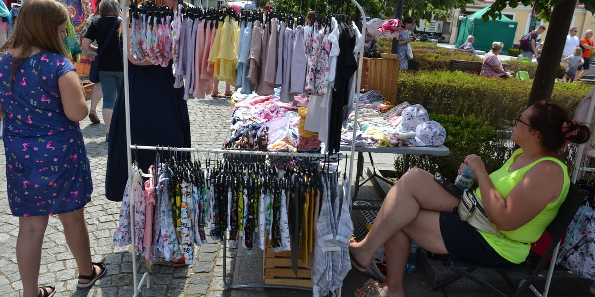 Po prawej stoi tyłem dziewczynka w niebieskiej sukience, w centrum stoi dwupoziomowy stojak z wiszącymi na wieszakach kolorowymi ubraniami, po prawej siedzi kobieta w jasnozielonej bluzce