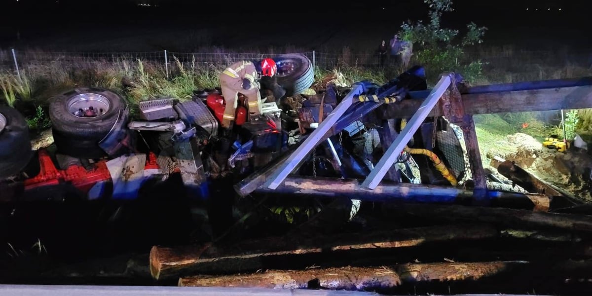 Rozbita ciężarówka, która brała udział w wypadku, leżąca w rowie. Widać też drewno i strażaka