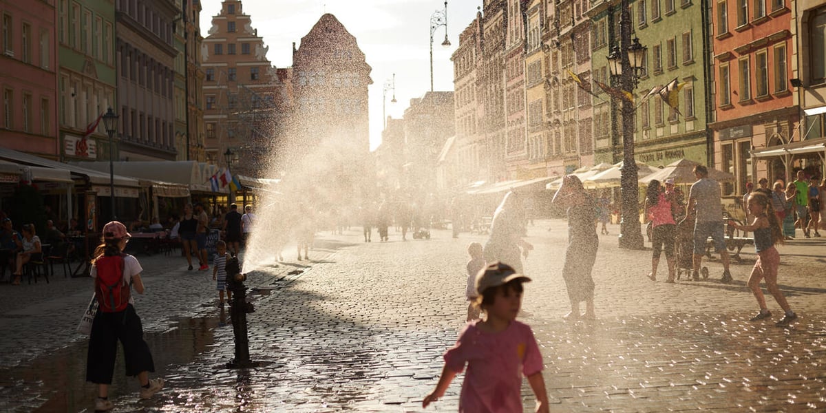 Rynek, brukowana ulica, z hydrantu leci woda, tworząc pióropusz kropel. Wokół biegają dzieci 
