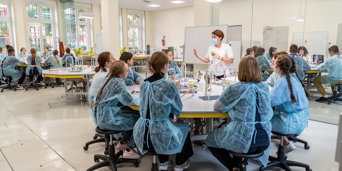 Sala warsztatowa Uniwersytetu Medycznego. Przy dużych wielokątnych stołach siedzi młodzież ubrana w błękitne ochronne fartuchy. W centrum stoi kobieta ubrana na biało, w masce ochronnej