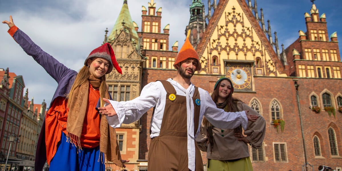 Festiwal Krasnoludków we Wrocławiu. Na zdjęciu trójka szczudlarzy w strojach krasnali, w tle wrocławski Ratusz