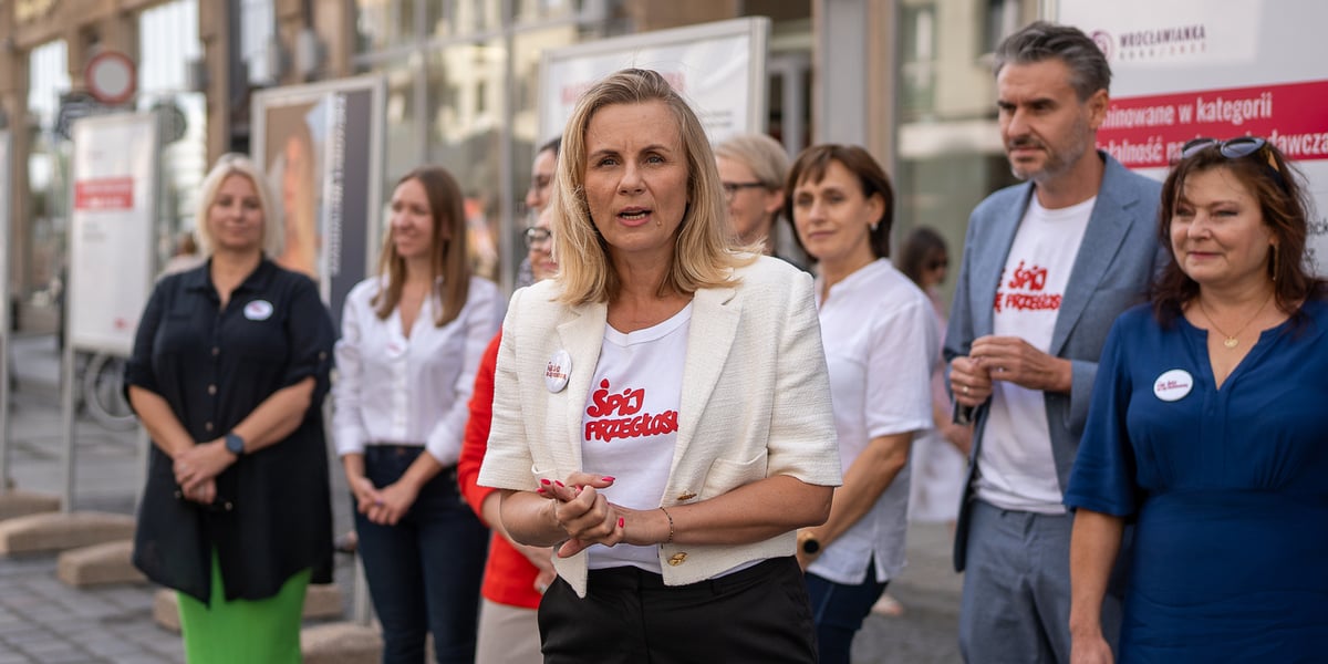 Kobieta w białym krótkim żakiecie stoi na tle stojących w rzędzie osób, z tyłu plansze; Joanna Nyczak podczas otwarcia wystawy Wrocławianka Roku 2022 i konferencji informującej o rozpoczęciu tegorocznej edycji plebiscytu  