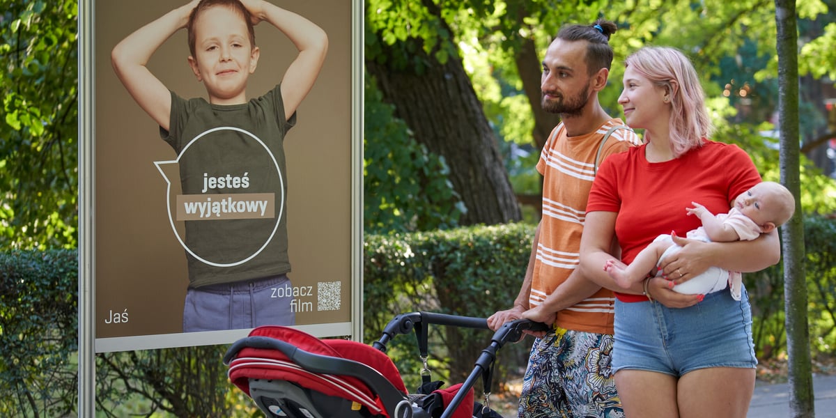 Rodzina, kobieta w czerwonej bluzce i spodenkach z niemowlakiem na rękach i mężczyzna w pomarańczowej koszuli w paski, pchający wózek, patrzą na planszę, na której widać chłopca. Napis na planszy: Czekam, aż do mnie powiesz: jesteś wyjątkowy. Jaś  