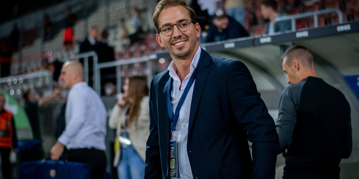 David Balda, dyrektor sportowy Śląska Wrocław, stoi uśmiechnięty przy ławce rezerwowych, z rękami w kieszeni, na nadgarstku ma zegarek.
