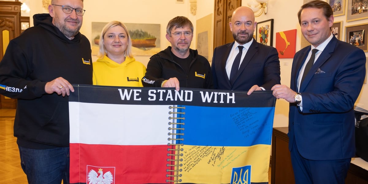 Prezydent Wrocławia Jacek Sutryk i delegacja ze Lwowa trzymają baner z flagami polską i ukraińską połączonymi szwem. Napis: We stand with Ukraine 