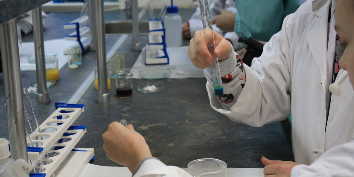 Stół w laboratorium, młode osoby ubrane w białe fartuchy trzymają w dłoni próbówki. Noc Biologów 2023