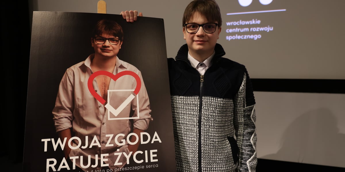 Szymon Konieczny, twarz kampanii „Zgoda to dar życia. Tak, chcę być dawcą”, przed plakatem z napisem „Twoja zgoda ratuje życie"