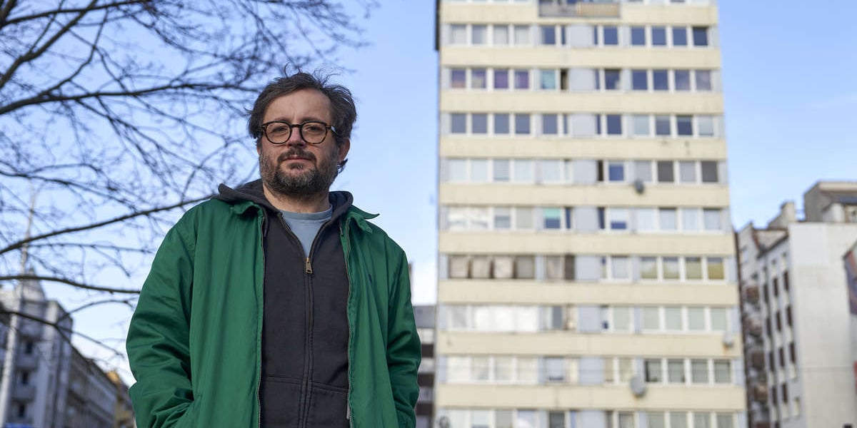 Mężczyzna w zielonej kurtce, na drugim planie wysoki budynek; dr hab. Łukasz Wojciechowski na tle Trzonolinowca 