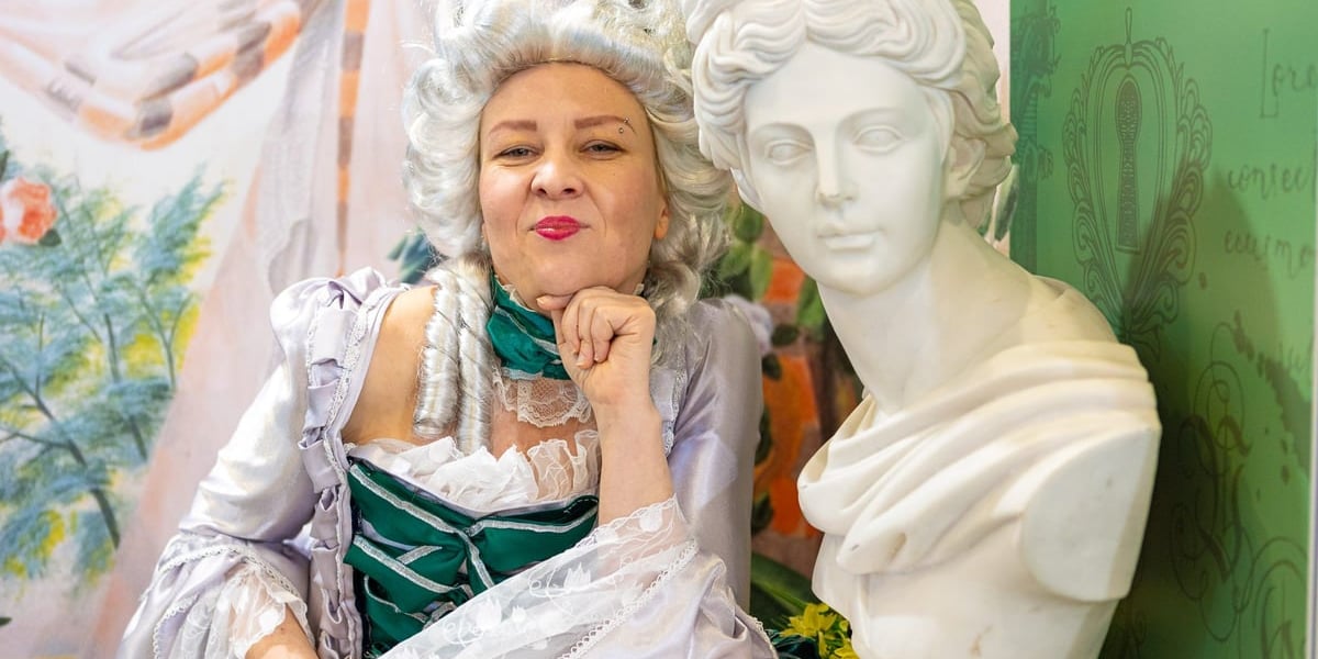 Uśmiechnięta kobieta w stroju z epoki w peruce, obok gipsowa figura kobiety